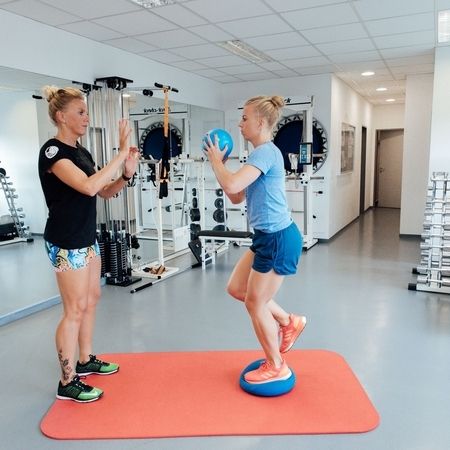 Ein Personal Trainer führt einen Kunden, der auf einer Übungsscheibe balanciert und einen Medizinball hält, in einem gut ausgestatteten Fitnessstudio.