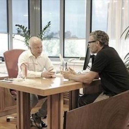 Zwei Männer unterhalten sich an einem Schreibtisch in einem hellen Büro, umgeben von Pflanzen und Büromöbeln.