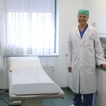 Ein Mediziner in Laborkittel und Mütze steht in einem hellen Klinikraum mit medizinischen Geräten und einem Untersuchungstisch für Patienten.