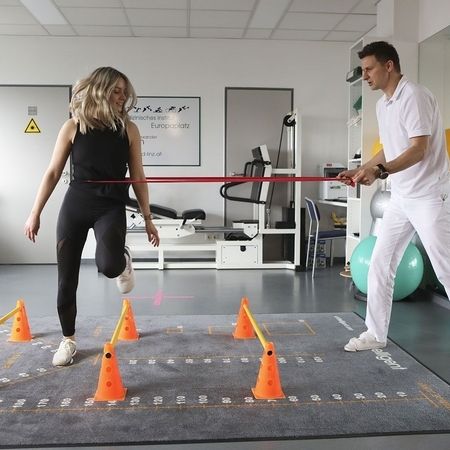 Eine Person führt eine Rehabilitationsübung mit einem Widerstandsband durch, während sie sich durch orangefarbene Kegel auf dem Boden eines Fitnessstudios bewegt.