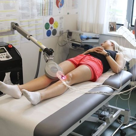 Eine Frau liegt auf einem Behandlungstisch mit einem Lasergerät auf dem Knie, das mit Kabeln verbunden ist, in einer klinischen Umgebung.